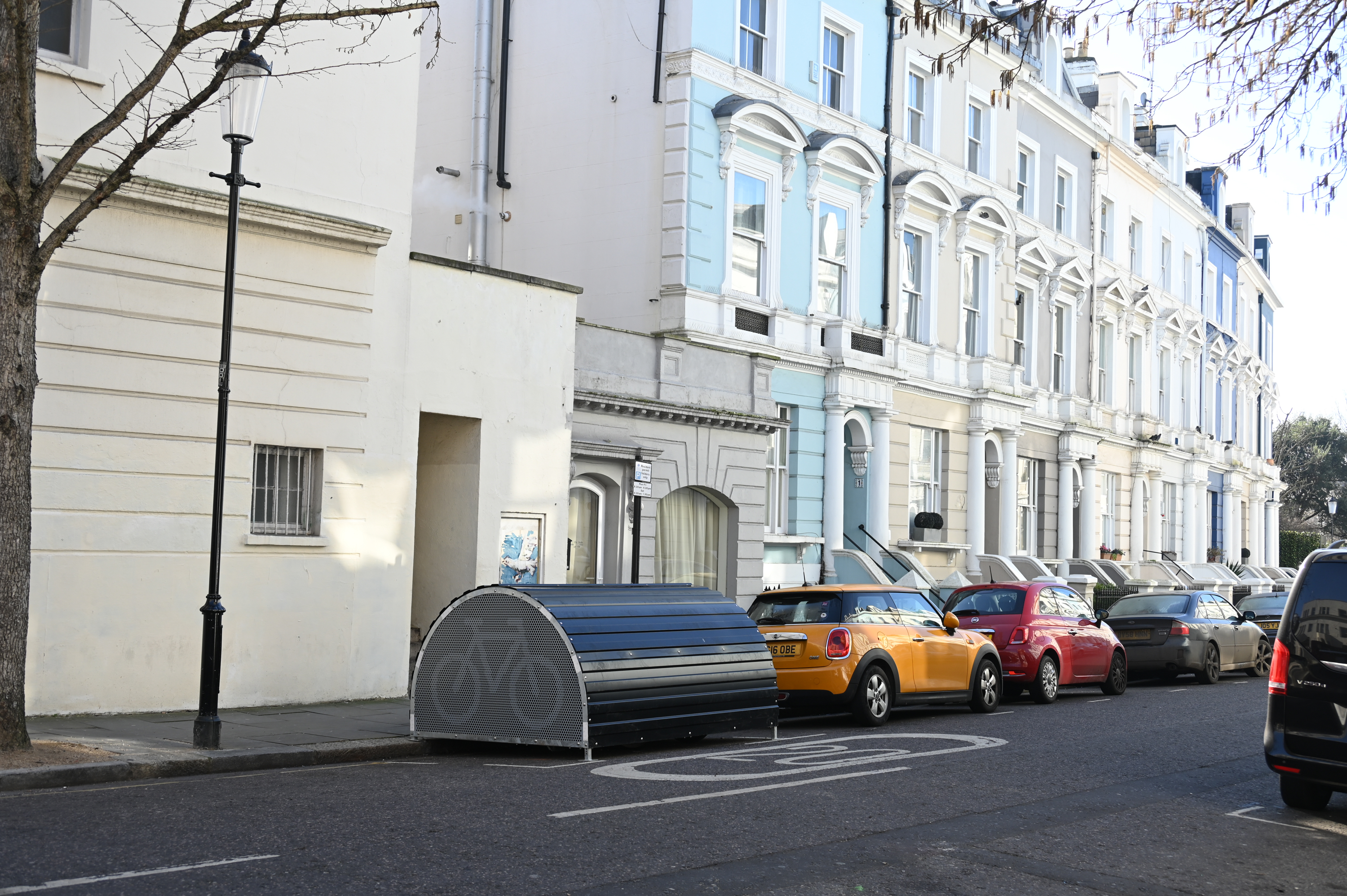 Example of a cycle hangar placed in the carriageway at the kerbside on a street.  The cycle hangar is shaped like a 'breadbin' opening on the footway side and has space for six bicycles within it.  