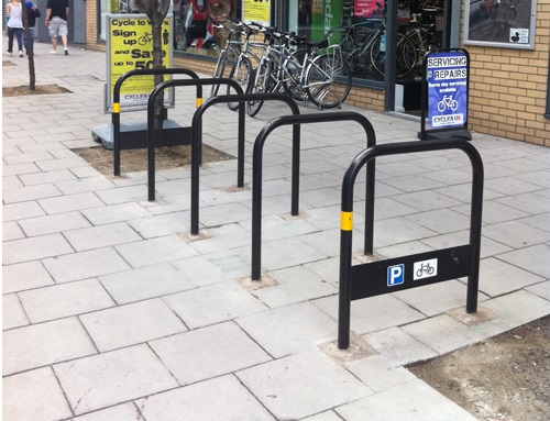 Photograph of five Sheffield cycle stands installed on footway between two trees, shops and pedestrians are in the background