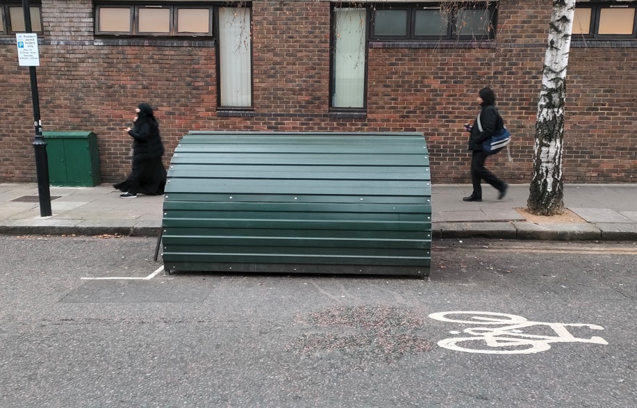 An example of a cycle hangar - a 'bread bin' type cycle storage unit in a dark forest green, installed within a parking bay.  It opens towards the footway. 