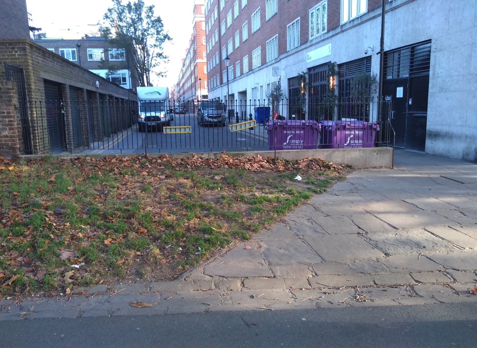 An image of the section of wall separating Phillimore Walk from Holland Walk. In front of the wall is a small section of grass and paving. Behind the wall is Phillimore Walk, buildings and large purple wheeled bins.